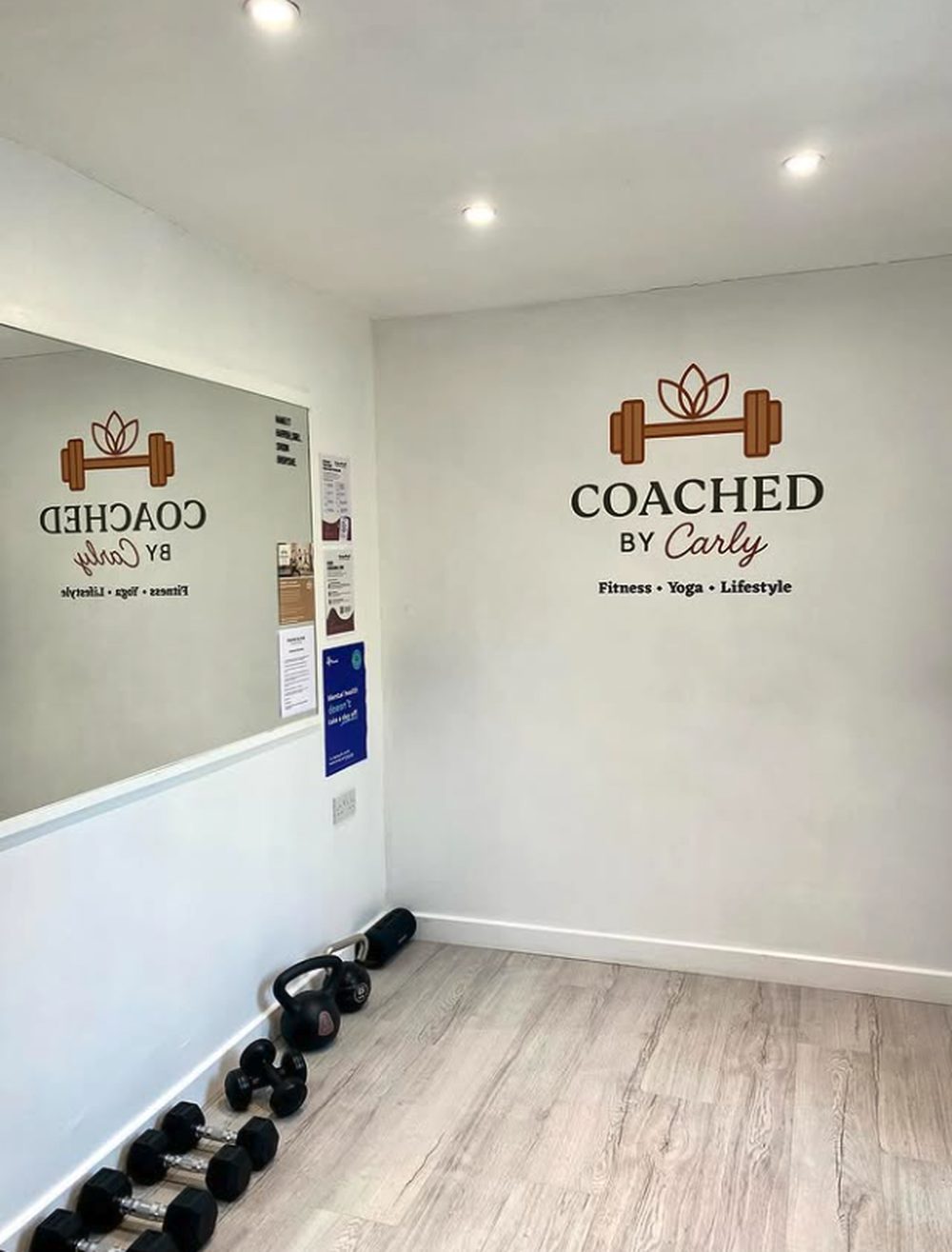 Carly's private studio in Balderton, with a long mirror, and dumbbells and kettlebells along the wall beneath the Coached by Carly logo.