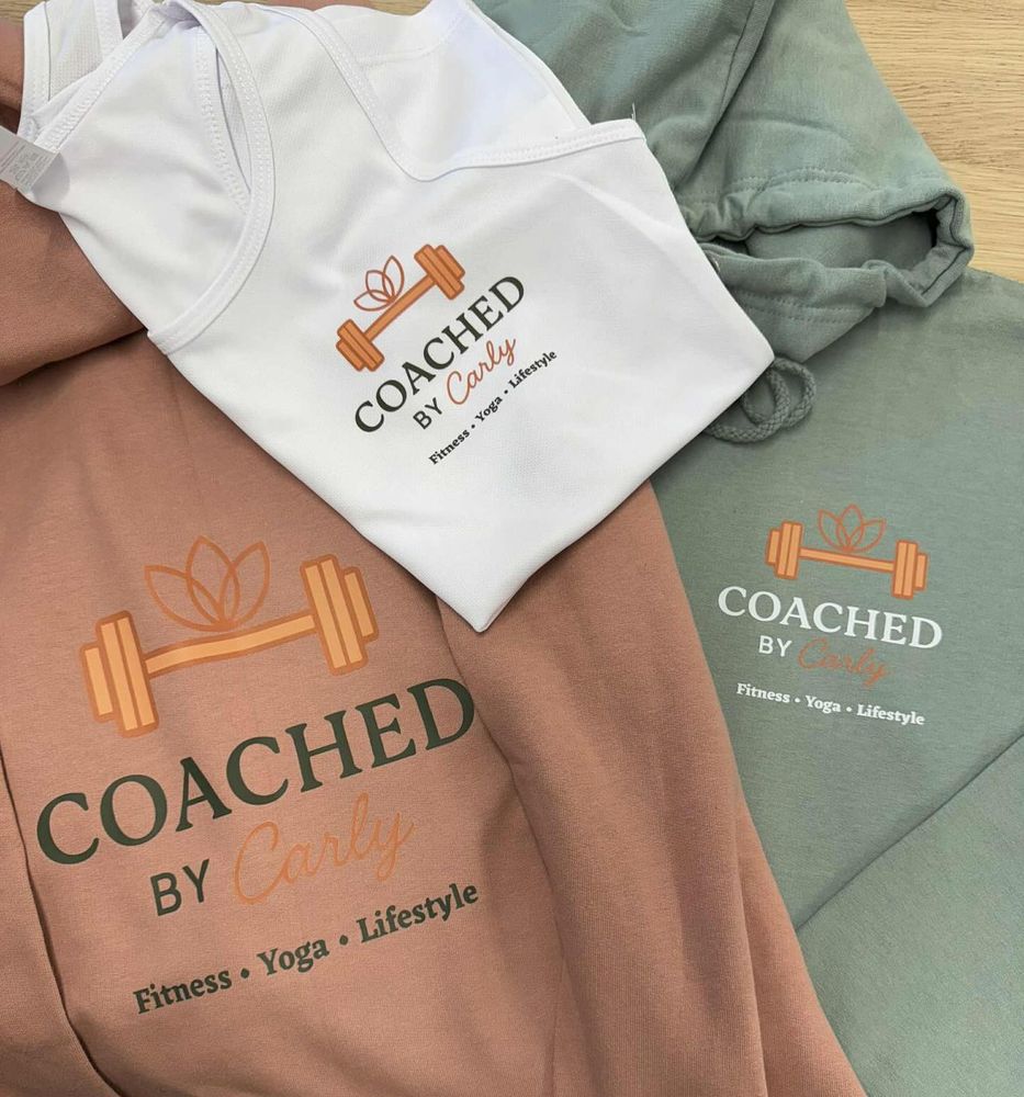 Coached by Carly clothing: a white vest, a rust sweatshirt and a sage sweatshirt, each printed with the dumbbell and lotus logo.